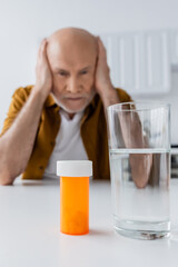 Glass of water and pills near blurred elderly man at home.