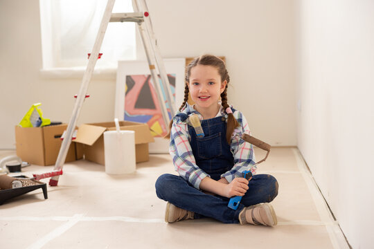 A very cute baby is sitting on the ground in a room under renovation. The girl is wearing denim overalls, a checkered shirt and beige sneakers. She holds a roller for painting walls in right hand.