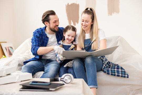 Happy family sitting on sofa in room where renovation work is taking place. Charming blonde woman helps husband daughter choose best shade of paint color to paint walls in living room.