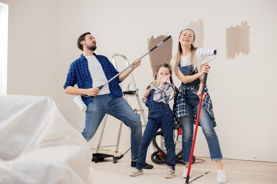 Cheerful family, dressed in denim, goofing around, having fun and rejoicing that renovations have begun in new house. They dance together with paint rollers and brushes loudly sing favorite song.