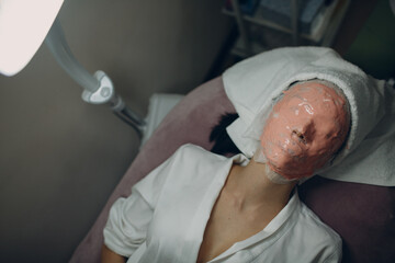 Alginate face mask application process. A female hands of a beautician holding spatula. Woman lying on a couch in office of cosmetologist. Facial skincare in the beauty spa salon.