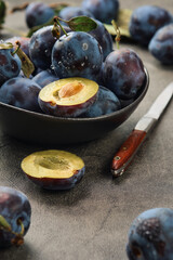 Selective focus, shallow depth of field. Ripe plums with leaves and halves of plums in a ceramic bowl on a gray table, next to a knife, close-up selective focus. Harvesting prunes in autumn.