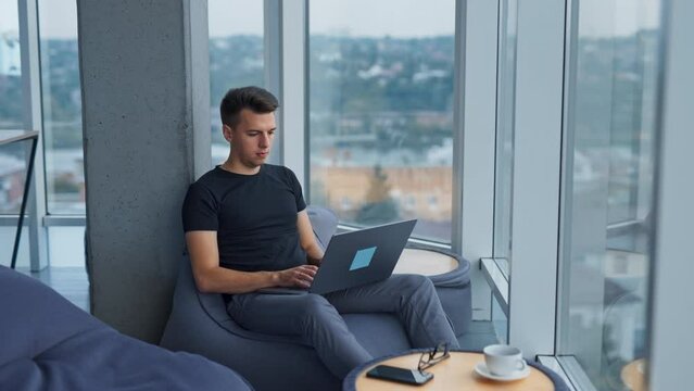 Dark-haired Caucasian man sitting in bean bag chair works on laptop. Male office employee at work. City at backdrop in blur.