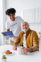 Smiling african american volunteer holding clipboard near elderly man with cup in kitchen.