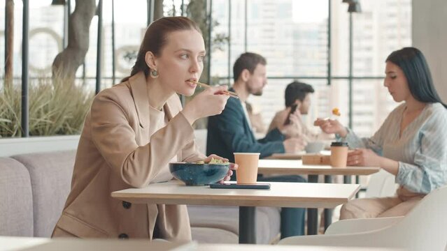Waist up slowmo of young Caucasian woman having Pan Asian food for lunch, sitting at table on food court with panoramic city view