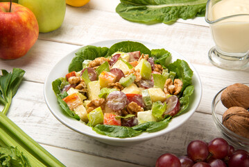 Waldorf salad of apples, celery stalks, walnuts, grapes, lettuce in a white salad bowl with ingredients on an old rustic white wooden table. Selected focus. - 524225033