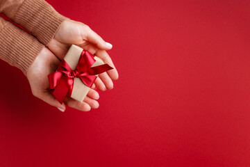 Female's hands holding gift box tied red bow on red background.