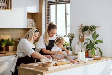 Excited family teaching girls kneading dough standing at table in modern light kitchen. Preschooler daughter helping holding knife cutting butter with mother help. Granny managing process.