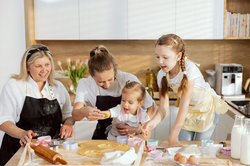 Young mother and pretty preschooler daughter pressing cookies with cookies cutter. Granny and older daughter watching managing process. Family having fun together in modern kitchen.