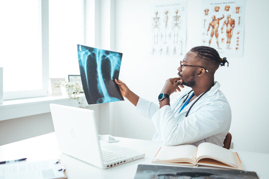 Healthcare, technology, rontgen, people and medicine concept - smiling male doctor in white coat with laptop computer looking at x-ray in medical office. Male doctor reviews x-ray