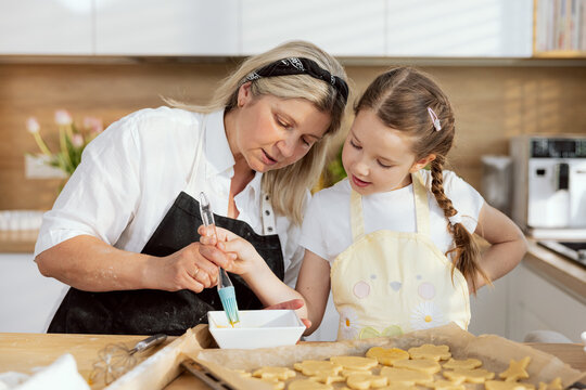 Young curious daughter having fun together with delighted happy mother. Cheerful mom teaching preparing dough before baking. Adorable kid holding silicone oil basting brush.