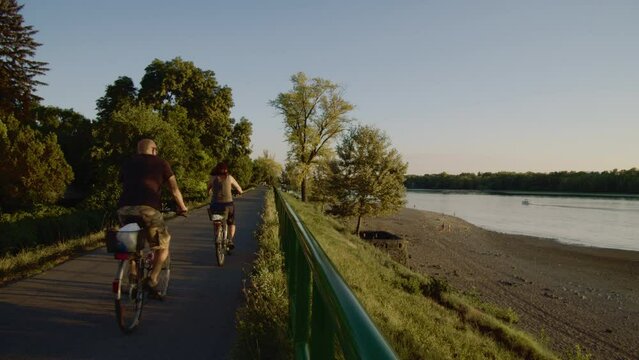Elderly man and woman are cycling on the cycle path. The bike path is located by the Danube. A motorboat travels on the Danube.