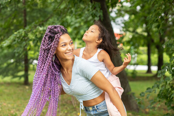 Fototapeta premium cute funny girl on the back of mom in the park, african woman playing with a child in the summer in nature, a happy family