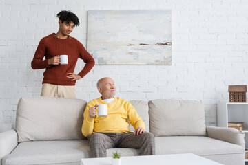 African american grandson holding cup near grandpa sitting on couch at home.