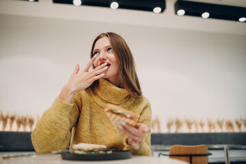Young european woman licks fingers after eating sandwich at indoor cafe. Health and diet.