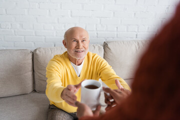 Blurred african american man holding cup of tea near smiling granddad on couch.