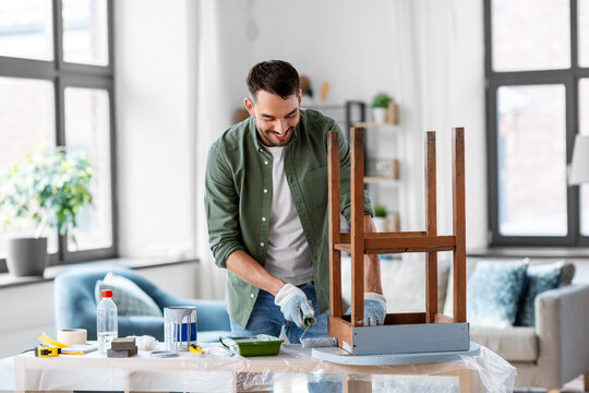 renovation, diy and home improvement concept - happy smiling man in gloves with paint roller painting old wooden table in grey color