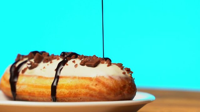 Sweet eclair with chocolate sprinkles, rotating on a wooden table on a blue background. Chocolate is poured on the eclair.