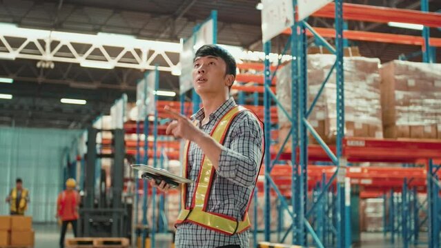 Young man Asian worker wearing helmet checking inventory and counting product on shelf in modern warehouse.