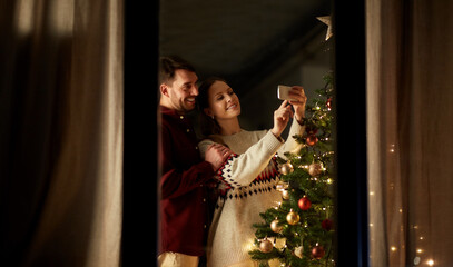 Fototapeta na wymiar winter holidays and people concept - happy couple with smartphone taking selfie at christmas tree at home
