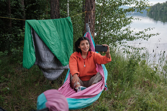 High angle view of modern young adult Hispanic woman sitting relaxed in hammock at riverside shooting video on smartphone for blog