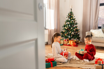 Obraz na płótnie Canvas christmas, winter holidays and childhood concept - happy girl and boy in pajamas opening gifts sitting on floor in front of each other at home