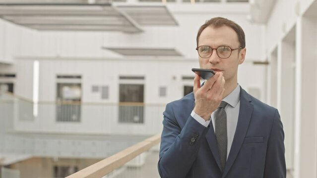 Waist up slowmo portrait of confident Caucasian businessman in blue formal suit, tie and eyeglasses recording voice message on smartphone then smiling at camera, standing at modern business center