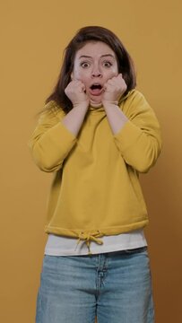 Portrait of woman being scared doing hand gesture touching face in fear feeling frightened and afraid over yellow background. Young person doing shocked face grimace being startled in studio.