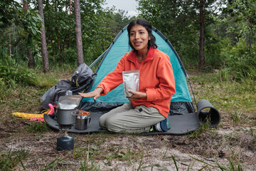 Young adult Hispanic woman telling what kind of food products she is using to cook meals while camping in mountain forest
