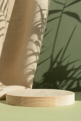 Wooden cylindrical podium, pedestal on a green background with beige curtain and a shadow of palm leaves. Scene for the presentation of a cosmetic product. Empty showcase