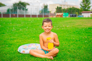 A cute smiling boy sits on the green grass in swimming trunks and holds a safe tanning product in his hands in a spray bottle