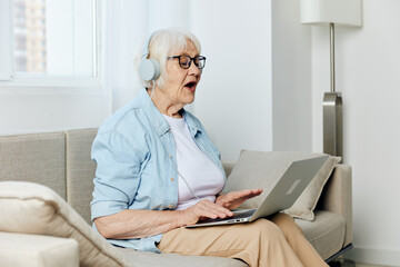a pleasant, happy elderly woman is sitting on a beige sofa and holding a laptop on her lap, communicating via video using headphones