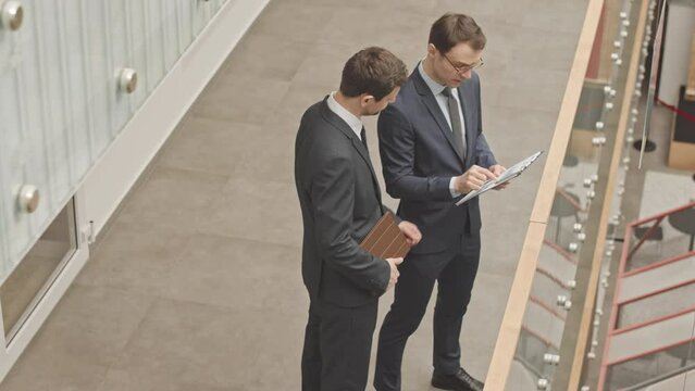 High angle slowmo of two smart businessmen in formalwear looking at printed financial charts and graphs on clipboard and having discussion, standing in corridor of modern business center
