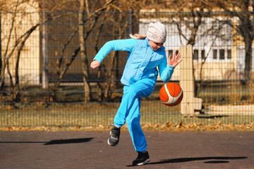 A child in blue outerwear 10 - 11 years old plays with a ball on a basketball court, outside.