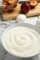 Fototapeta na wymiar Bowl with flour on white marble table, closeup. Cooking of delicious plum cake
