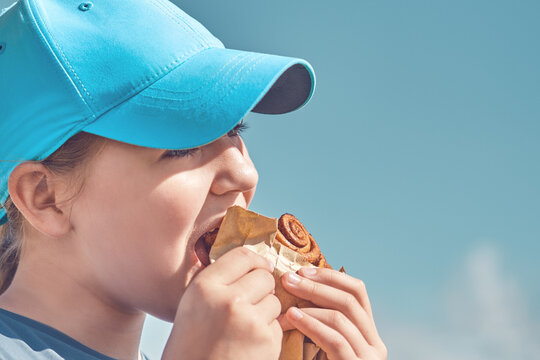 A Teenager In A Cap Eats Pastries Against A Blue Sky On A Summer Day Outside. High School Girl Eating Cinnamon Bun, Face Close Up.