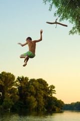 Boy jumping into the river from the swinging rope in summer - 524250055