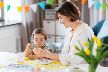 Obraz premium holidays, family and people concept - happy mother and little baby daughter coloring easter eggs with watercolors at home