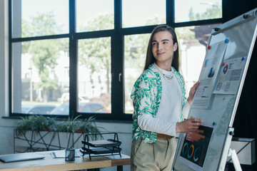 Smiling queer designer standing near graphs on flip chart in office. - obrazy, fototapety, plakaty
