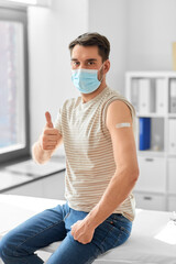 health, medicine and pandemic concept - male patient in mask with patch on his arm after vaccination showing thumbs up gesture at hospital