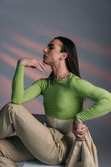 Side view of trendy queer model holding jacket while sitting on chair on abstract background.