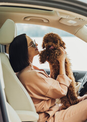 Young brunette woman is a happy owner of redhead dog toy poodle, adult girl sitting inside car with her four paws pet