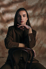 Queer person in jacket looking at camera while sitting on chair on abstract brown background.