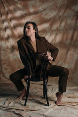 Barefoot queer person in blazer sitting on chair on abstract brown background.