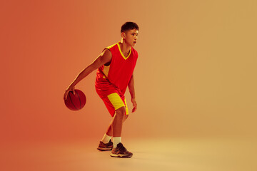 Fototapeta na wymiar Portrait of young man, basketball player training isolated over orange studio background in neon light. Dribbling exercises