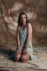 Young tattooed queer person looking at camera while sitting on abstract brown background.