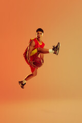 Fototapeta na wymiar Portrait of young man, basketball player in motion, jumping isolated over orange studio background in neon light