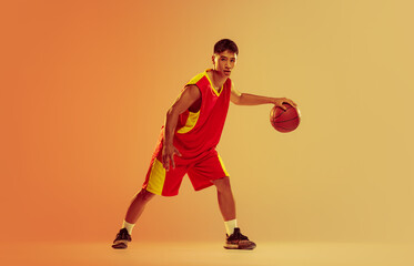 Portrait of young man, basketball player training isolated over orange studio background in neon...