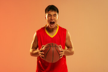 Portrait of young man, basketball player screaming in emotions isolated over orange studio background in neon light. WInning game