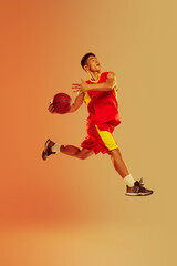 Dynamic portrait of young man, basketball player throwing ball into basket isolated over orange...